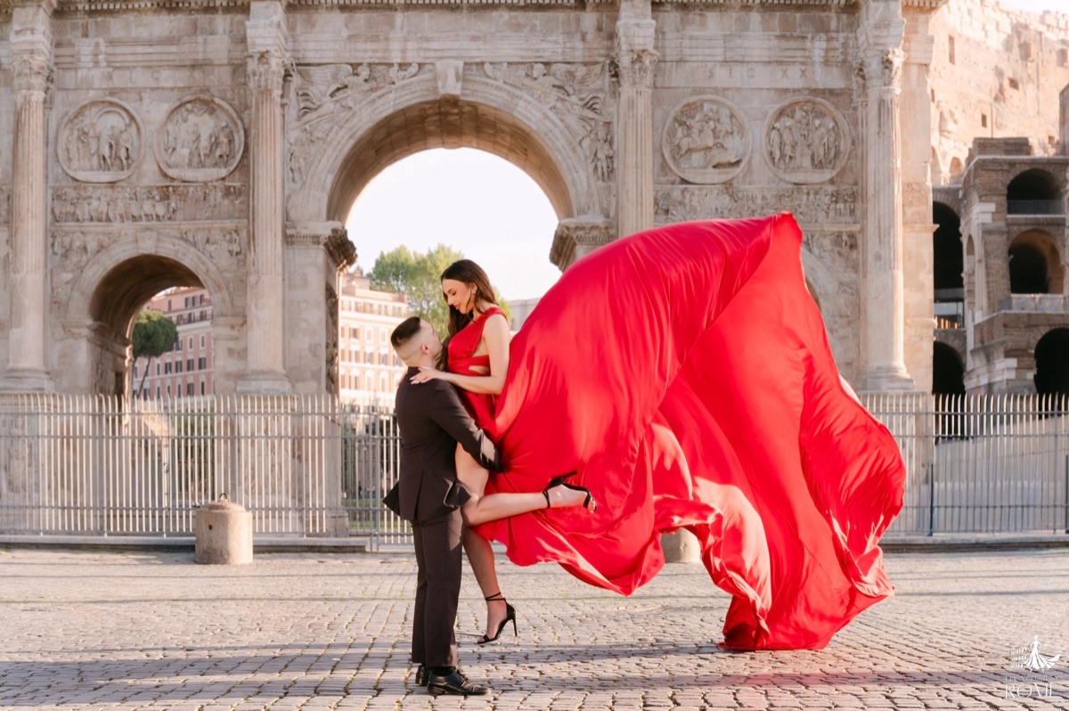 Posa flying dress di coppia The Dip: lui la inclina all'indietro sostenendola, strascico rosso che vola, Arco di Costantino, Roma