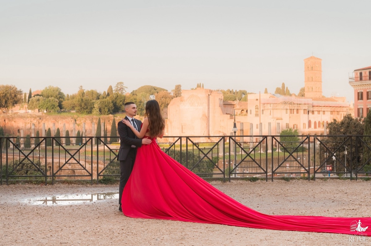 Posa flying dress di coppia The View: coppia ferma quasi abbracciata, lei di spalle, strascico rosso disteso a terra, Fori Imperiali, Roma