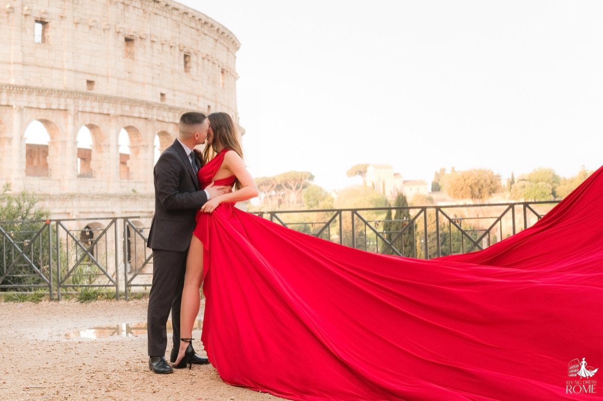 Posa flying dress di coppia The Embrace: lui la tiene per la vita, strascico rosso lanciato dietro, Colosseo all'alba, Roma